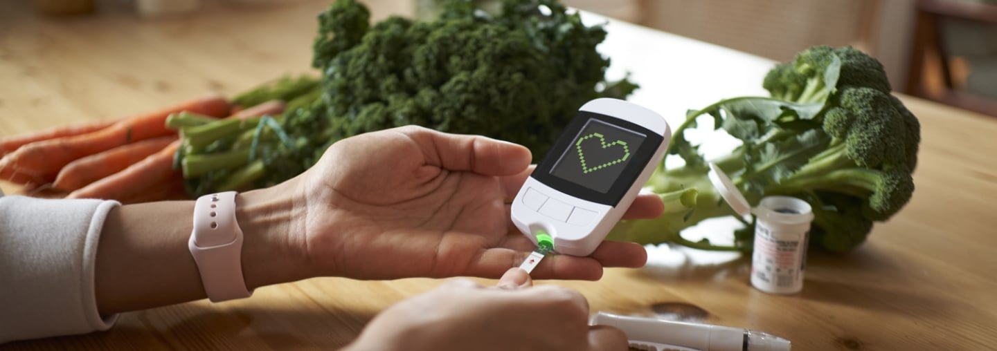 A closeup of a woman using a glucose meter on a table with broccoli and carrots in the background